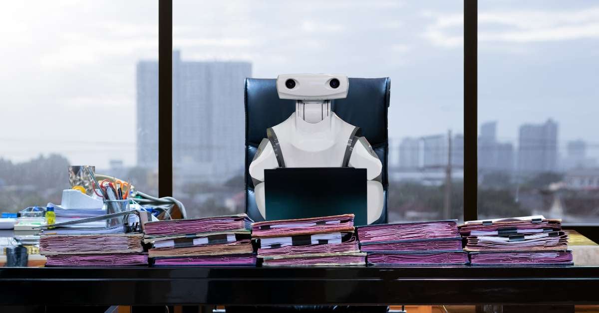 A robot sits at a desk working on a laptop to autonomously file work documents.